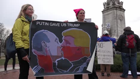 Dos-Manifestantes-Políticas-Femeninas-Llevan-Un-Cartel-De-Donald-Trump-Y-Vladmir-Encerrados-En-Un-Abrazo,-Washington-Dc