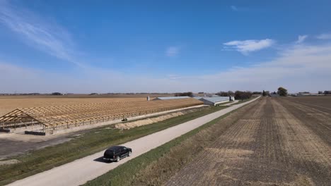 Aerial-Drone-Footage-Of-A-Black-Suv-On-A-Farm-Road-Passing-Farm-Buildings-Under-The-Blue-Skies-Of-Rural,-Midwestern-Iowa