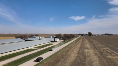 Drohnenaufnahmen-Aus-Der-Luft-Eines-Schwarzen-Geländewagens-Auf-Einer-Landstraße,-Die-An-Landwirtschaftlichen-Gebäuden-Vorbeiführt,-Unter-Dem-Blauen-Himmel-Des-Ländlichen-Iowa-Im-Mittleren-Westen