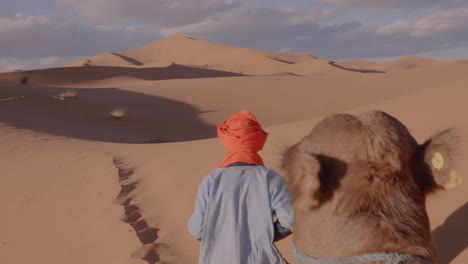 POV-riding-a-camel-into-the-Sahara-desert,-guided-by-man-in-turban