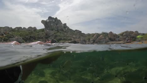 Fpv-half-underwater-pov-of-male-legs-relaxing-while-floating-on-sea-water-with-fish-swimming-and-rocks-in-background-at-Cala-Della-Chiesa-cove-on-Lavezzi-island-in-Corsica,-France