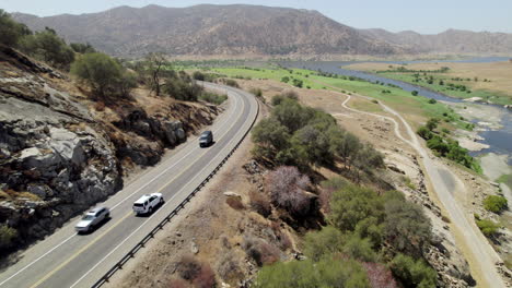 Aerial-View-Of-White-SUV-Driving-Along-Hillside-Road-Towards-Desert-Landscape-In-California