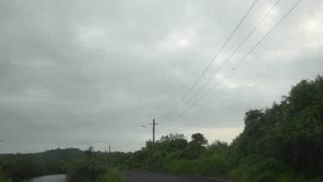 POV-shot-of-a-vehicle-travelling-along-an-asphalt-road-with-an-overhead-powerline-running-along-the-roadside-on-a-grey-gloomy-overcast-day,-Panjim,-India