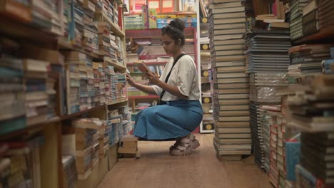 An-attractive-Asian-female-reading-the-blurb-on-the-back-of-a-hardcover-book-in-the-isle-of-a-bookstore,-India