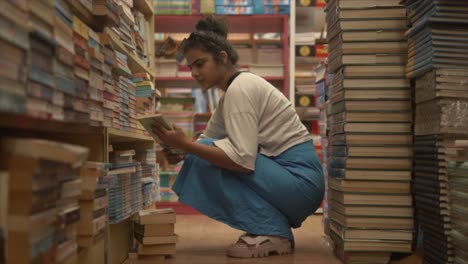 A-beautiful-female-reading-the-cover-and-title-of-a-book-while-crouched-on-her-haunches-in-the-middle-of-an-isle-in-a-bookstore,-India