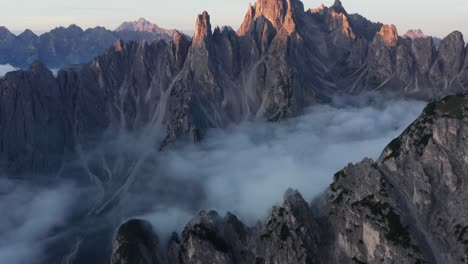 Drone-tilt-up-reveals-craggy-Cadini-di-Misurina-peaks-lit-up-by-sunrise-with-cloud-inversion-in-valley---cinematic-views-in-Dolomite-Alps,-Italy