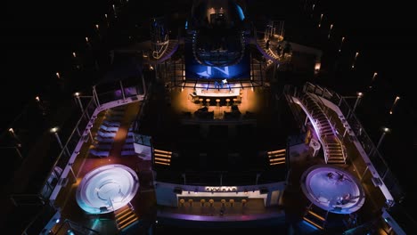 Aerial-drone-top-down-shot-over-blue-top-deck-pool-facilities-and-jacuzzi-over-huge-cruise-liner-at-night-time