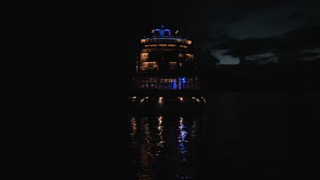 Shot-of-back-of-Luxury-cruise-ship-sailing-along-the-bay-past-the-coastline-on-a-dark-night