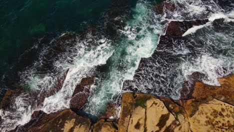 Top-view-pan-of-multiple-blue-green-ocean-waves-smashing-against-rocky-coastline-in-Australia