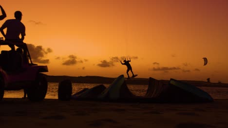 Slomo-shot-at-twilight-of-kitesurfer-jumping-in-front-of-friends-sitting-on-buggy---dark-silhouettes-against-orange-sunset-sky