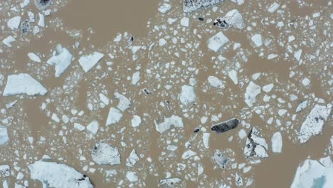 Top-down-aerial---brown-water-with-pieces-of-floating-ice-in-glacial-lake