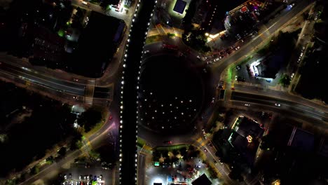 Aerial-top-down-view-of-big-large-traffic-circle-with-bridge-over-it-at-night-with-the-lights-of-a-big-city
