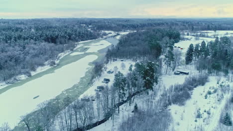 Aerial-drone-panning-shot-over-cottages-beside-frozen-river-and-forest-along-winter-landscape-on-a-cloudy-day