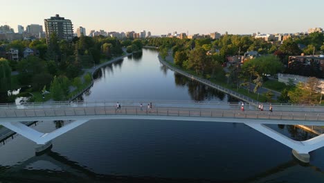 Aerial-View-Of-Pedestrians-Walking-Across-Flora-Footbridge-Over-Calm-Reflective-Rideau-Canal-In-Ottawa