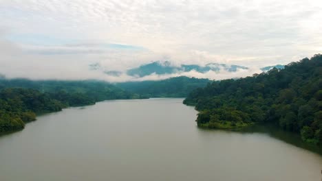 Volando-Sobre-El-Agua-Turbia-Del-Lago-Con-Vistas-A-La-Montaña-Envuelta-Por-Nubes-En-El-Fondo-En-Semenyih,-Malasia
