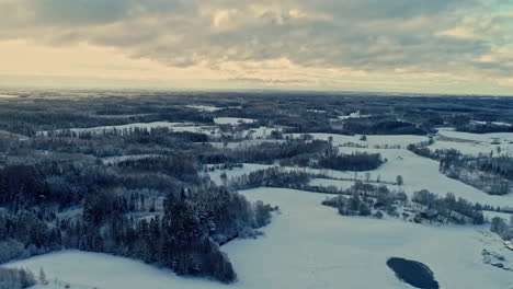 Beautiful-drone-aerial-shot-over-white-snow-covered-rural-landscape-with-frozen-pond-on-a-cloudy-day