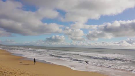 Ein-Einsamer-Surfer-Steht-Und-Beobachtet-Die-Wellen,-Den-Strand-Und-Den-Teilweise-Bewölkten-Himmel-Im-Hintergrund
