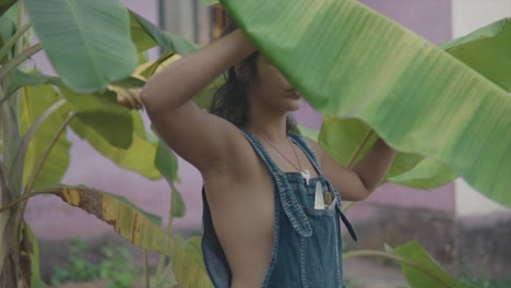 Medium-handheld-shot-of-a-beautiful-indian-model-dressed-in-a-blue-jean-dress,-chain-and-a-tightening-hair-elastic-on-her-arm-lifting-a-large-leaf-of-a-plant-in-the-garden