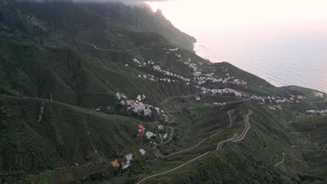Beautiful-aerial-view-of-several-towns-between-the-green-mountains-of-Tenerife-linked-by-paved-roads-with-the-sea-in-the-background-at-sunset