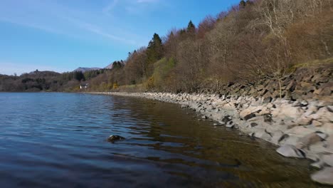 Low-fly-along-waters-edge-on-Loch-Lomond-shore-next-to-rocky-beach-on-a-sunny-day