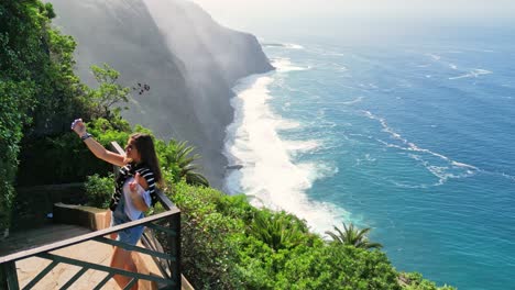 A-young-tourist-woman-takes-pictures-with-her-mobile-phone-at-the-viewpoint-of-a-cliff-overlooking-the-ocean-in-Tenerife,-Canary-Islands
