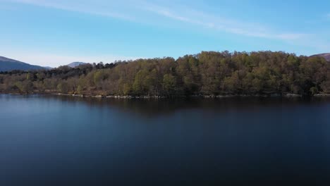 Low-fly-along-water-of-Loch-Lomond-and-lift-up-over-island-to-reveal-Ben-Lomond-and-mountains-on-a-sunny-day