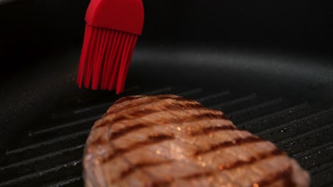 Close-up-handheld-dolly-shot-of-a-seared-beef-steak-in-a-hot-pan-being-coated-with-oil-from-a-red-brush-for-a-delicious-dinner