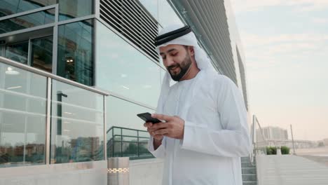 Hombre-árabe-Feliz-En-Kandoora-Usando-Un-Dispositivo-De-Teléfono-Celular-Emiratí-árabe-Local-En-Una-Ubicación-Comercial-Con-Un-Edificio-De-Vidrio-Como-Fondo