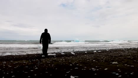 Hombre-Caminando-En-La-Playa-De-Arena-Negra-En-La-Laguna-Glaciar-En-Islandia