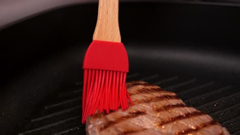 Close-up-shot-of-a-red-oil-brush-with-wooden-bamboo-handle-brushing-a-steak-in-the-hot-pan-with-oil-while-frying-for-a-delicious-dinner