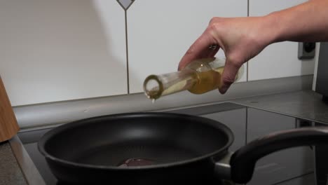 Medium-handheld-shot-of-a-woman's-hand-with-a-bottle-filled-with-oil-while-she-adds-high-quality-sunflower-oil-to-a-hot-pan-for-cooking-in-the-kitchen-for-a-delicious-dinner