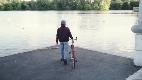Man-is-walking-towards-the-lake-with-his-bike-in-his-hand,-peaceful-and-calm-atmosphere