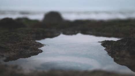 Rockpool-reflection-shows-cloudy-sky-rack-focus-waves-lapping-at-shoreline