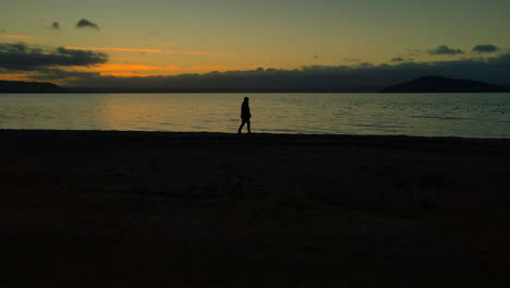 Slow-pan-to-a-Lonely-Person-at-Dusk-slowly-walking-alone-along-shore-at-sunset-with-horizon-in-background