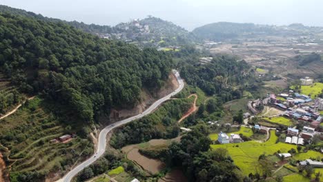 Eine-Wunderschöne-Luftaufnahme-Der-Vorberge-Des-Himalaya-Mit-Einer-Straße,-Die-Sich-Entlang-Des-Fußes-Eines-Hügels-Mit-Verstreuten-Feldern-Und-Häusern-Windet
