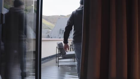 Man-in-a-grey-hoody-and-beanie-comes-into-hotel-room-dropping-off-his-travel-backpack-onto-a-chair,-opening-the-door-and-going-on-to-the-rooms-balkony-terrace-with-a-views-of-europes-winery-mountains