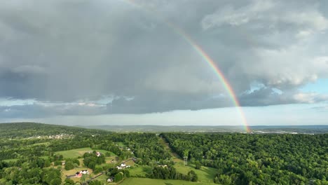 An-aerial-flight-over-the-green-rural-farmland-after-a-storm-with-the-clouds-and-a-rainbow-in-the-sky