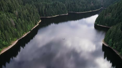 Aerial-of-boat-float-on-calm-lake-with-cloud-reflection-sided-by-forest