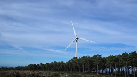 Distant-shot-of-single-wind-turbine-spinning-with-few-clouds-behind-and-pine-forest-below
