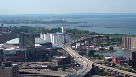 An-aerial-view-of-the-city-of-Buffalo,-New-York-and-its-infrastructure-of-bridges-and-buildings-beside-Lake-Erie-with-some-wind-turbines-in-the-background
