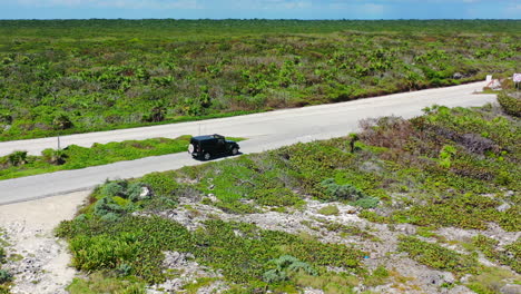 SUV-driving-on-empty-island-road-full-of-lush-greenery-in-Cozumel-Mexico,-aerial