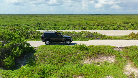 Jeep-driving-on-lush-green-tropical-island-on-sunny-summer-day-in-Cozumel-Mexico,-aerial