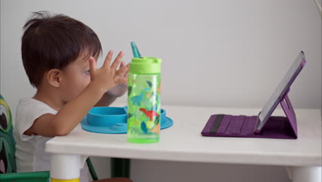Young-latin-baby-boy-eating-his-breakfast-on-a-children-table-watching-a-show-on-a-purple-tablet