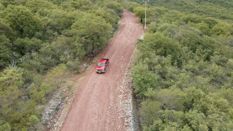 Aerial-view-of-a-red-pickup-truck-driving-on-a-muddy-road-in-a-mountain-forest,-tracking-shot