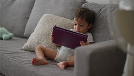 Cute-young-latin-baby-boy-in-a-light-blue-baby-romper-sitting-on-a-grey-couch-watching-cartoons-on-a-purple-tablet