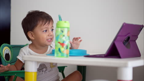 Small-latin-toddler-having-fun-watching-a-show-on-a-purple-tablet-while-having-breakfast-on-a-white-small-table-with-colorful-legs