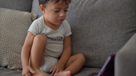 Nettes-Lateinamerikanisches-Kleinkind-In-Einem-Hellblauen-Babyspielanzug,-Das-Auf-Einer-Grauen-Couch-Sitzt-Und-Cartoons-Auf-Einem-Lila-Tablet-Anschaut