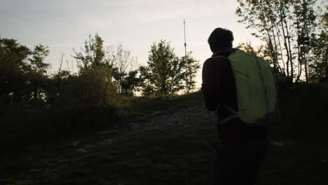 Hiker-with-a-green-backpack-walks-up-a-hill-towards-the-sun,-camera-tracking-from-behind