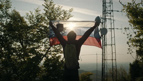 Man-holding-Slovenian-flag-towards-the-summer-sun-near-trees-and-a-radio-tower-on-peak-of-mountain-Slivnica