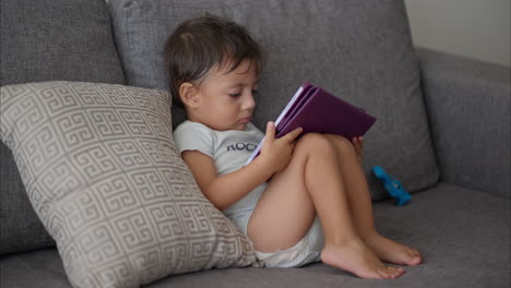 Junger-Kleiner-Lateinamerikanischer-Junge-In-Einem-Hellblauen-Babyspielanzug,-Der-Auf-Einer-Grauen-Couch-Sitzt-Und-Einen-Film-Auf-Einem-Violetten-Tablet-Genießt